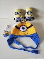 Photo shows one handmade Minion hat with blue edges and one eye, braided ties, and three Minion figurines in a row: one smaller and two larger figures.