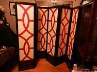 Shows two black wooden 3-panel room dividers with red and white geometric designs on cream background, standing partially open indoors.
