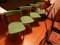 Four green wooden accent chairs arranged in a row, front view showing seats and legs on hardwood floor.