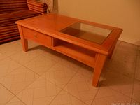 Front-side view of the tan wood coffee table showing the glass inset, drawer, and lower shelf, placed on tiled floor.