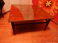 Square mid-century modern coffee table with brown wood frame and glass top, set on hardwood floor, photographed from angled overhead to show table surface and leg design.