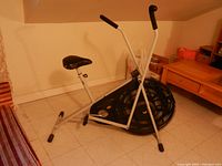 Side view of the white framed exercise bike showing the fan wheel, black adjustable seat, and handle bars with foam grips.