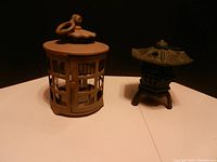 Two green metal garden lanterns displayed on a white surface against dark background, showing circular lantern with latch and pagoda votive holder.