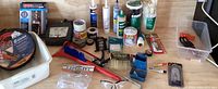Wide shot showing the full collection of garage and useful essentials including jumper cables, mesh screen door, tape, caulking tubes, safety goggles, and various tools and accessories arranged on a wooden surface.