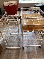 Two white metal storage units with wire mesh baskets and drawers, one on wheels, with a red rubber tub and white wastebasket on top.
