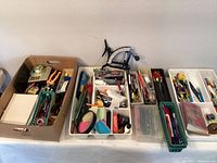 Photo showing various pens, pencils, scissors, cutters, small office tools, and trays organized in cardboard and plastic containers.