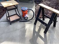 Three items on driveway: sawhorse, work table, and orange shop vac with black hose and extra filter beside it