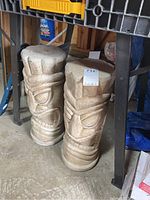 Pair of cement Tiki head style garden sculptures standing on concrete floor under workbench, showing front carved facial features and wood grain texture tops