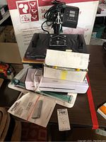 Heat press machine on top with digital display, red and yellow pressing surfaces. Below it is an Amazon Basics thermal laminator, white, with power cord coiled on top. Various packing materials underneath.