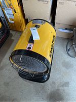 Front and side top view of yellow cylindrical heater sitting on concrete floor surrounded by boxes.