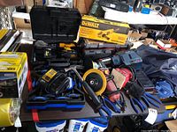 Overview of table with DeWalt drill in open case, batteries, charger, DeWalt grinder box, Kobalt hex key set, red Sanders, pliers and hammer