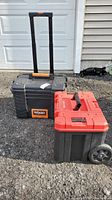Photo of two large plastic toolboxes, Ridgid with extension handle and wheels, and Craftsman with red lid, placed outdoors on gravel