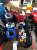 Photo showing various rolls of tape, quick connect hose kits, and black plastic tool case on a table with Milwaukee saw box partially visible.