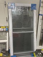 Front view of a vinyl double hung window, showing the white frame and glass panes, wrapped in plastic with a label.