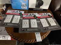 Three unopened packages of Feit Electric USB wall outlets placed on a round wooden table.