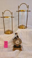 Photo showing pair of brass and glass hurricanes with clear cylindrical glass and brass handles, alongside a decorative jester clock with gold and black colors.