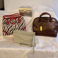 Four purses displayed on white fabric background: brown faux crocodile leather handbag, grey leather shoulder bag, faux zebra print handbag with red trim, silver metal lunchpail handbag.