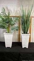 Two faux plants side by side in tall square whitewashed wooden pots on black surface against white wall.