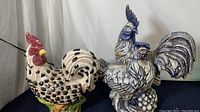 Two ceramic bird figurines displayed on black cloth against white backdrop. One is a hand-painted spotted chicken with red comb, the other a blue and white detailed rooster.