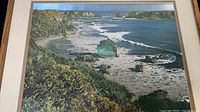 Framed lithograph showing a coastal beach scene with ocean waves and rocky outcrop. The metallic accents on the rocks and waves are visible. Frame has lightly worn edges.
