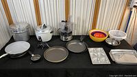 Full view of kitchen items including Hamilton Beach food processor, cookware, bakeware, mixing bowls and utensils displayed on black tablecloth.