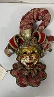 Front view of handmade Joker Venetian mask with detailed colorful face, ornate headdress in red and gold fabric, and ruffled collar