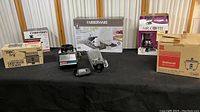 Wide shot of all appliances displayed on a black table against a neutral background, showing boxed and unboxed items including a deep fryer, slow cooker, coffee maker, rice cooker, blender, and kitchen scale.