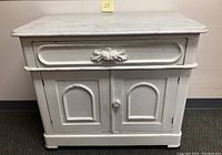 Full front view of the antique white painted commode showing marble top, drawer, and cabinet doors.