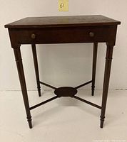 Front view of antique wooden side/end table with turned legs, drawer with two brass knobs, and cross stretcher base.