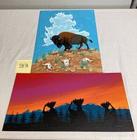 Two acrylic paintings displayed on a table: one showing a bison on a hill with skulls and clouds, the other showing Native American figures in silhouette against a sunset.