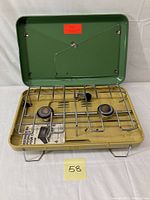 Open 2 burner camping stove in green and yellow/beige metal with metal grate and burners visible.