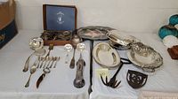 Wide view of silverplated serving utensils, Doll's Lemon Server set in box, serving trays, telephone handset cover, wrought iron rake, and cast iron trivet on table.