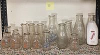 Overview of all 16 vintage milk bottles arranged in a row on floral tablecloth showing various sizes and branding.