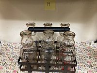 All six vintage Aquidneck Island Creamery milk bottles placed in a black metal delivery rack on a floral fabric background.