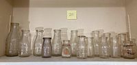 Full view of all 15 clear glass vintage milk bottles lined up together on a shelf with varying sizes and shapes