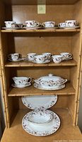 Full view of multiple shelves of china including 13 tea cups and saucers, covered casserole dish, platters, and bowls with floral and gold trim design.