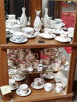 View of a wooden cabinet shelf holding numerous porcelain items including teacups, saucers, and small vases with floral and butterfly designs all arranged neatly.
