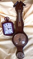 Wooden wall-mounted barometer with thermometer and a small purple clock on a beige fabric background.