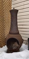 Full view of rusted metal chiminea with mesh door and tall chimney against lattice fence and snow-covered ground.