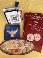 Canadian commemorative items displayed including a metal tin with a Canadian stamp in original packaging, a set of millennium coins in an oval display case, and Warman's coin collecting folders for cents and 25 cents coins.