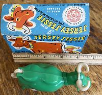 Top-down view of green plastic Jersey Jessie cow toy with horns, face and attached white tail, along with original illustrated box behind it. A ruler below shows measurement around 5 inches.