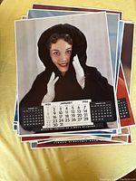 Stack of large 1956 calendar pages with front page showing January featuring a woman in a black hooded coat, calendar dates clearly visible at bottom