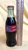 Image of unopened Coca-Cola bottle standing next to a ruler measuring about 7.5 inches tall showing the commemorative label.