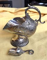 Side view of silver plated sugar cube server showing floral embossing, ornate handle and pedestal base with matching scoop