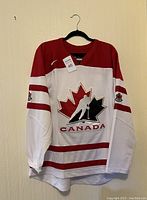 Front view of white and red Team Canada hockey jersey with large maple leaf logo and player silhouette on chest.