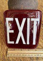 Front view of rectangular red glass EXIT sign with white lettering and a wooden ruler for size reference, showing approximately 8-9 inches tall.