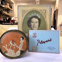 Three vintage tins displayed upright: large Queen Elizabeth II rectangular tin, light blue Player's Navy Cut Cigarettes rectangular tin, and round orange Three Flowers Dusting Powder tin with stylized women illustration.