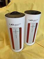 Two UE Boom 360 wireless speakers standing upright on a yellow background, showing the white body with red mesh sides and black top with hanging loop.