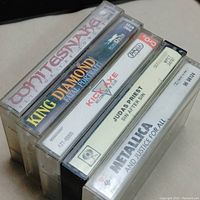 Five cassette tapes stacked showing spines with band names and album titles: Whitesnake, King Diamond Fatal Portrait, Kick Axe Welcome to the Club, Judas Priest Sin After Sin, Metallica ...And Justice for All.