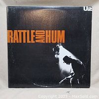 Front cover view of U2 Rattle and Hum album with black background, orange title text, and silhouette of band member playing guitar.